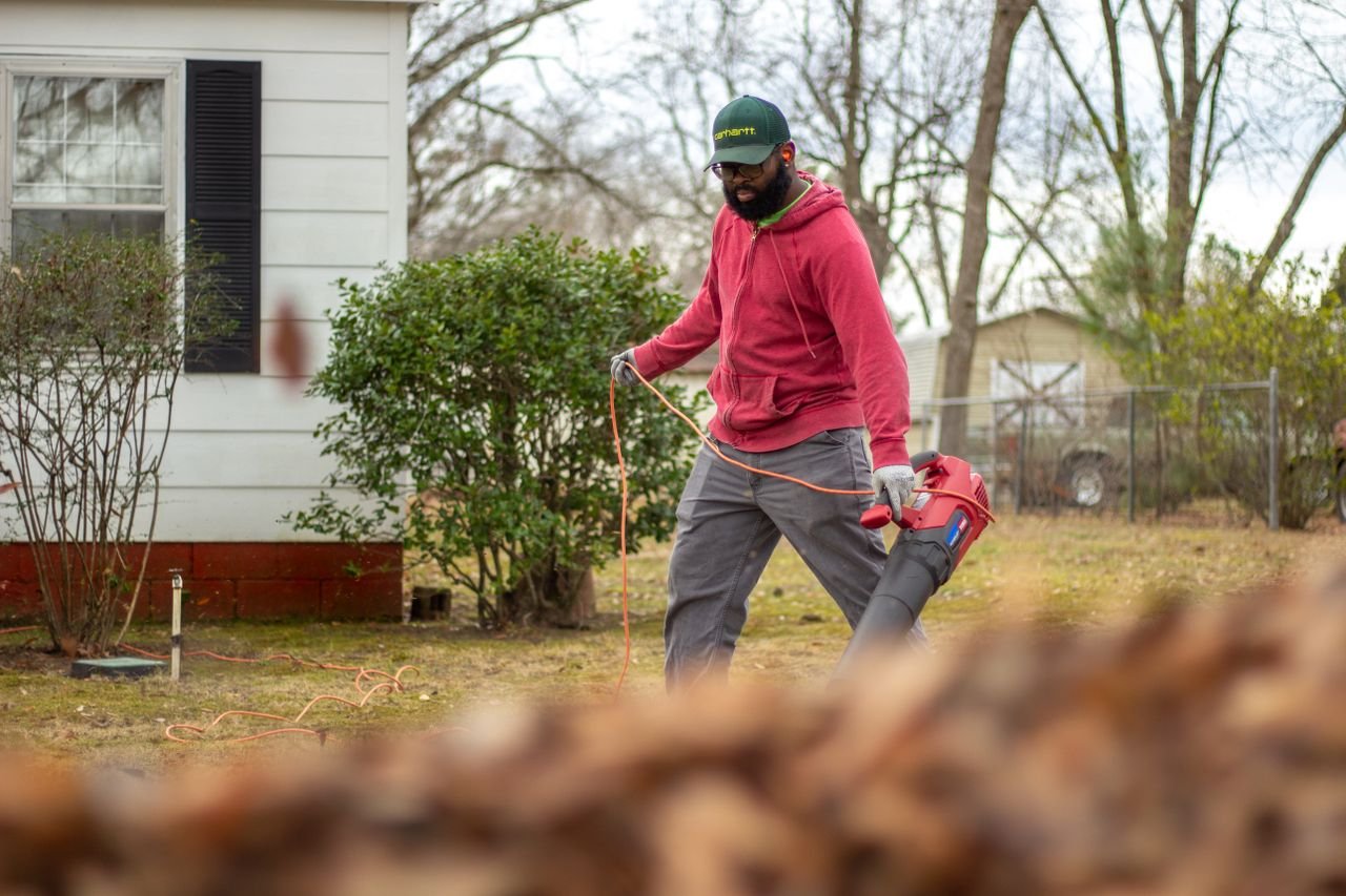 Home A man in a red jacket operates a leaf blower in an autumn backyard, clearing leaves.